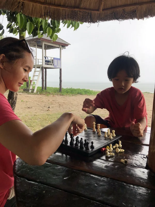 An adult and child playing chess at a shaded table beside the sea