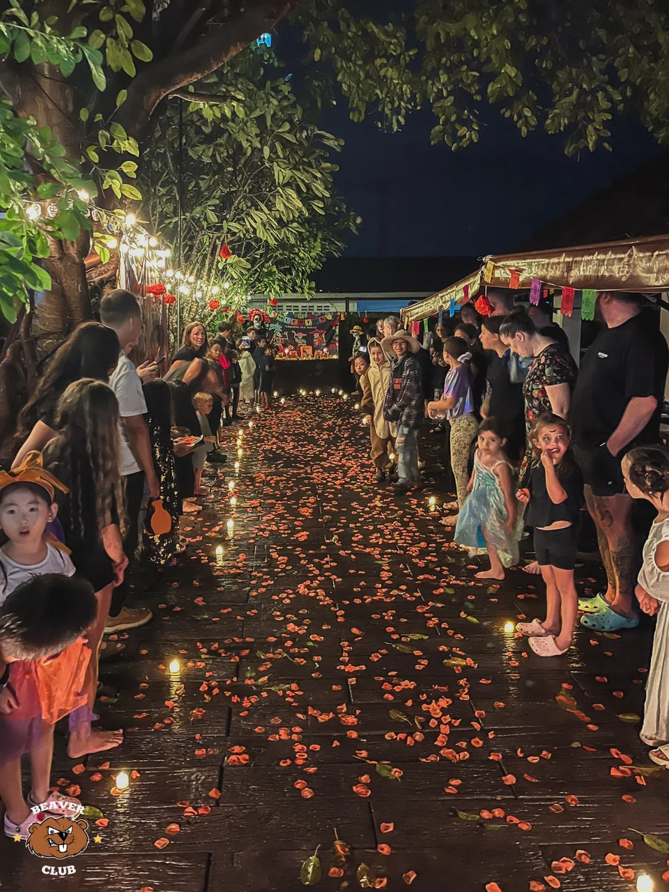 Families gathering along a petal-covered walkway beneath outdoor lights at a Halloween celebration