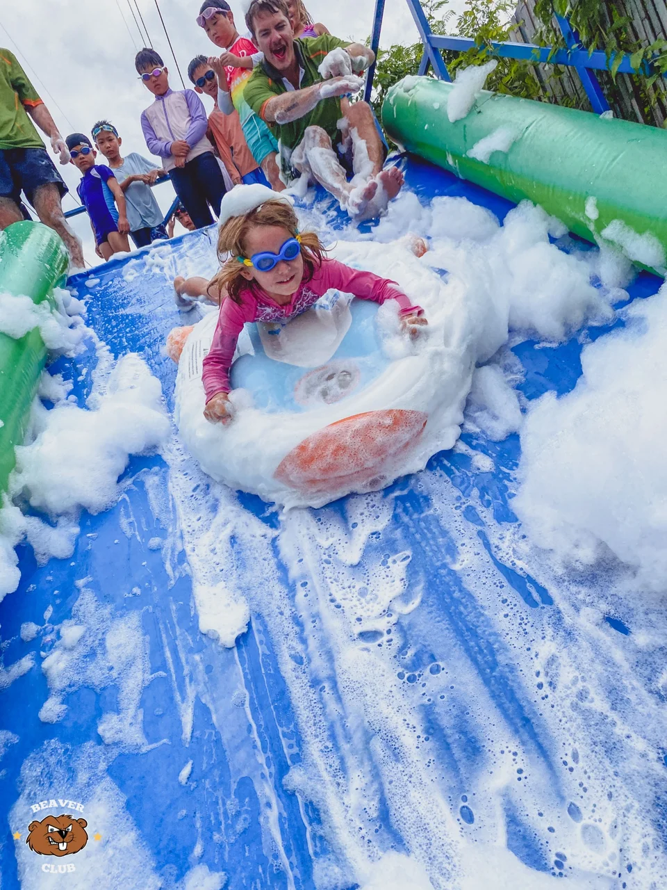 A child sliding through foam on the blue inflatable water slide while friends wait behind